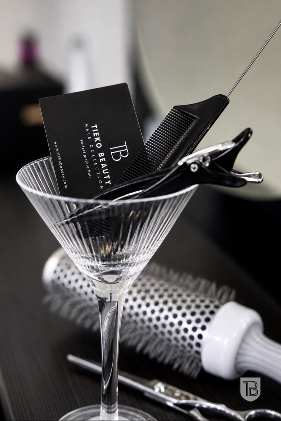 Tieko Beauty business card, comb and sectioning clip in a martini glass beside a round brush