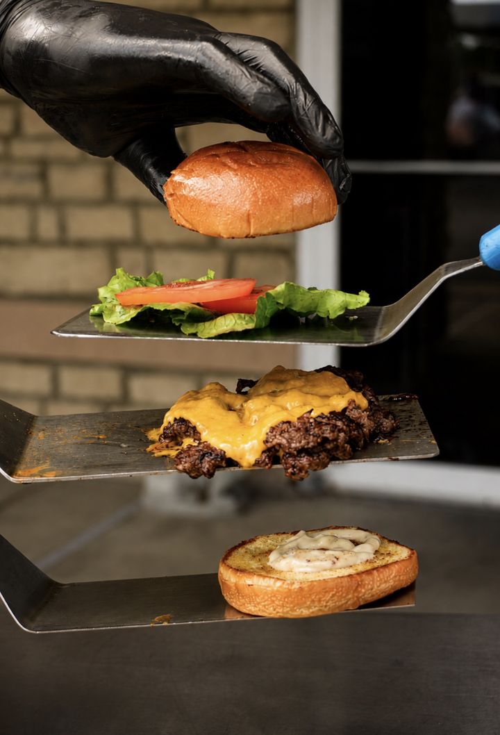 Gloved hand stacking a smash burger layer by layer on spatulas