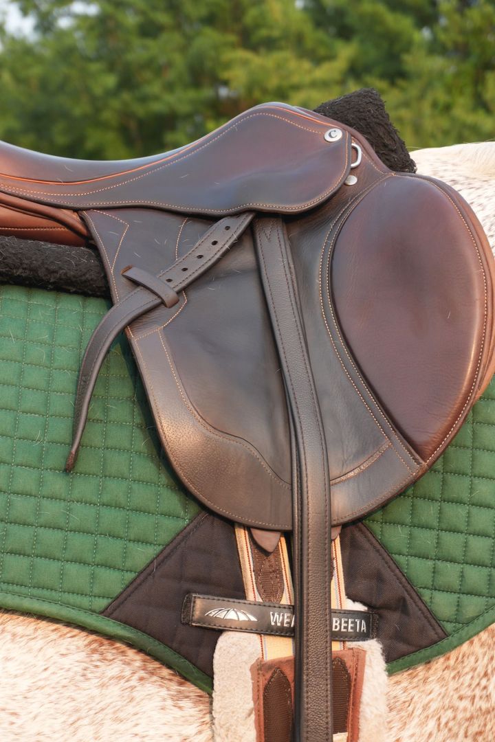 Close-up of a brown leather saddle with orange stitching on a green quilted pad and a speckled grey horse